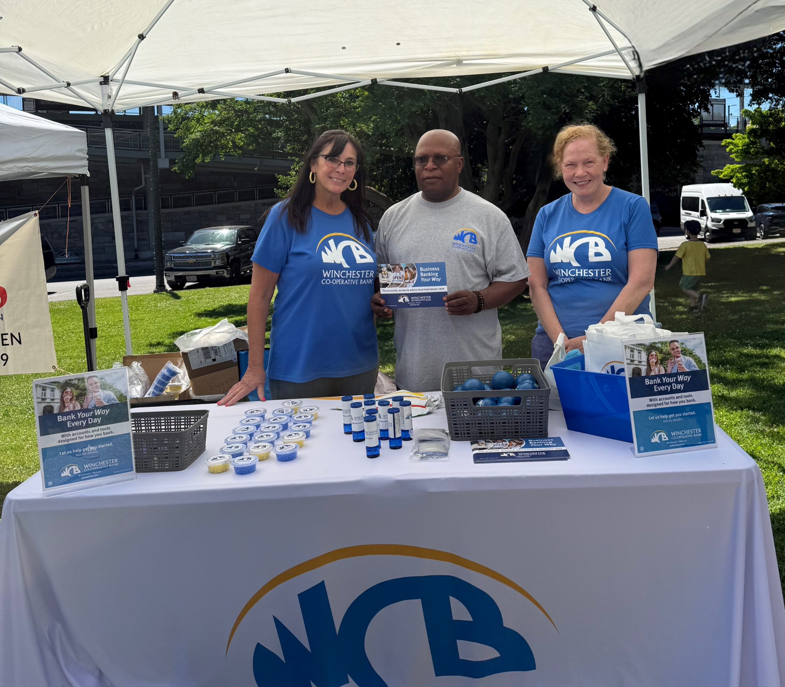 Karen, Sam and Julie in their blue and grey WCB tsheets standing at the table with the WCB tablecloth